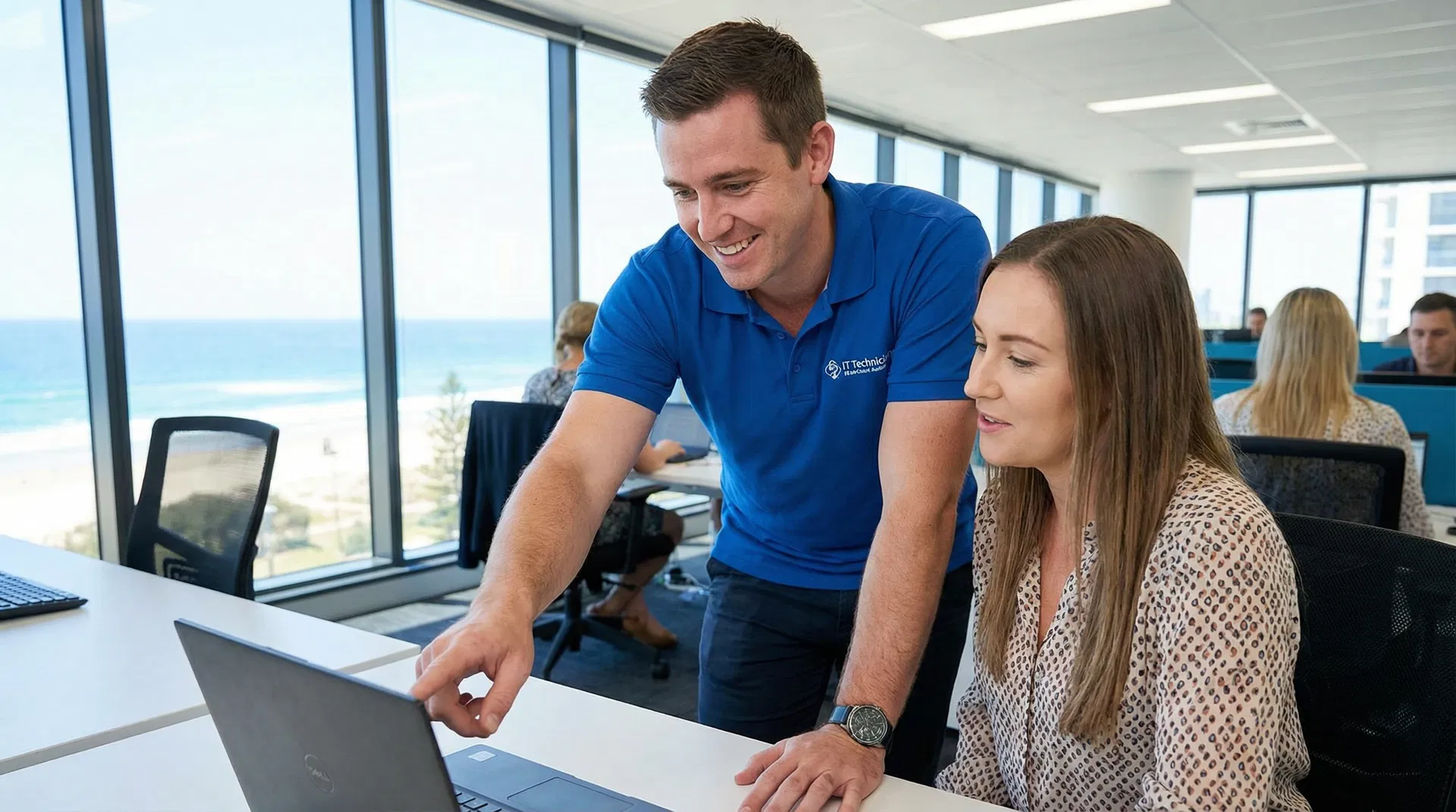 A friendly IT technician in a polo shirt helping a staff member at their desk in a sunny Gold Coast office